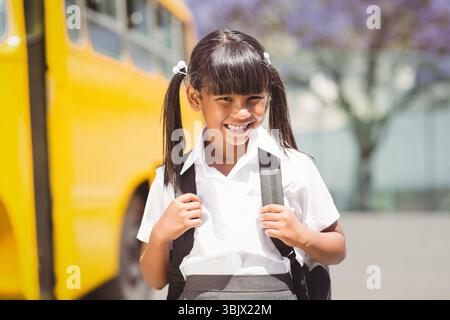 Bambina in uniforme scolastica in piedi sul marciapiede di scuolabus giallo, con zaino nero Foto Stock