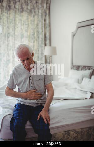 Uomo anziano seduto sul bordo di un letto non fatto in camera da letto con luce soffusa e tenda floreale e lampada Foto Stock