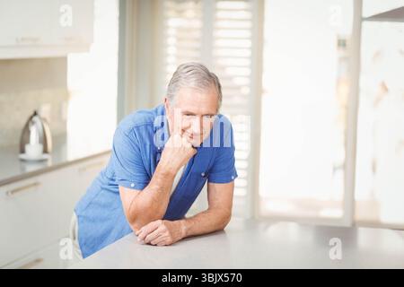Uomo anziano appoggiato al piano di quarzo in una cucina moderna con bollitore e persiane Foto Stock