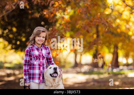 Un bambino bianco in piedi accanto al Golden retriever nel parco autunnale illuminato dal sole, che tiene un cane che accarezza il guinzaglio nero Foto Stock