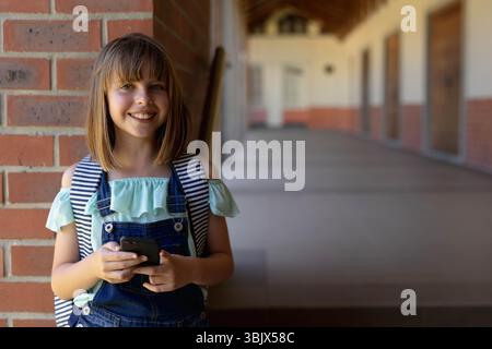 Studentessa bambina sorridente e con lo smartphone in mano sul passaggio pedonale del campus, copia dello spazio Foto Stock