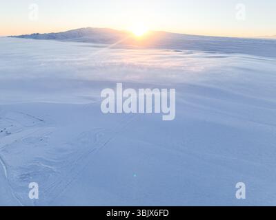 Una vista aerea di un tranquillo paesaggio innevato con il sole che sorge all'orizzonte, che emette un caldo bagliore Foto Stock