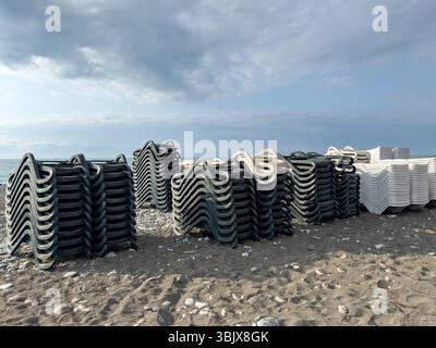 Lettini sdraio impilati su una spiaggia rocciosa sotto il cielo nuvoloso alla fine della stagione estiva. Concetto di pausa turistica, fuori stagione, gestione costiera. Niente persone. Foto Stock