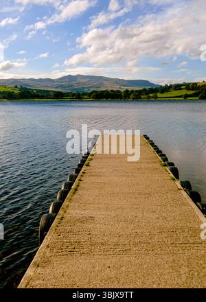 Un molo in cemento si estende in un lago, con montagne e un cielo nuvoloso sullo sfondo. Foto Stock