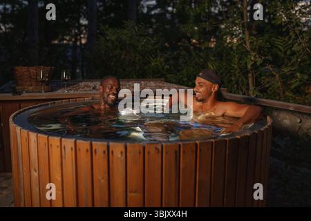 Coppia gay gay gay gay gay afroamericana che si rilassa in una vasca idromassaggio privata all'aperto di notte, godendosi il lusso di una spa glamping bevendo gustoso vino rosso. Foto Stock
