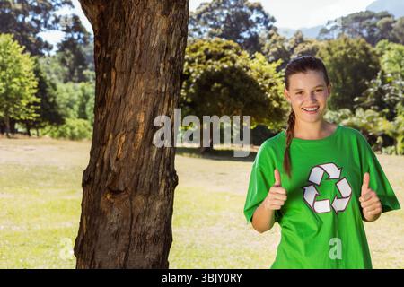 Donna in piedi accanto al tronco dell'albero in un prato soleggiato che indossa una camicia verde riciclabile e uno spazio per le copie Foto Stock