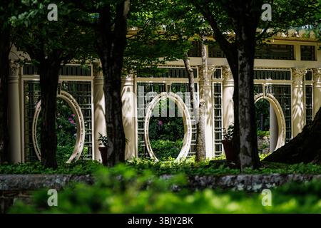 Elegant garden pavilion with circular archways seen through trees, dappled sunlight, lush foliage, peaceful estate garden, architectural detail. Foto Stock