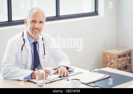 Il medico senior scrive appunti alla reception nella sala consultiva con stetoscopio, tazza da caffè e appunti Foto Stock