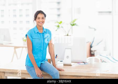 Donna seduta su una scrivania di legno in un luminoso ufficio moderno, con monitor per computer, pianta e tazza da caffè Foto Stock
