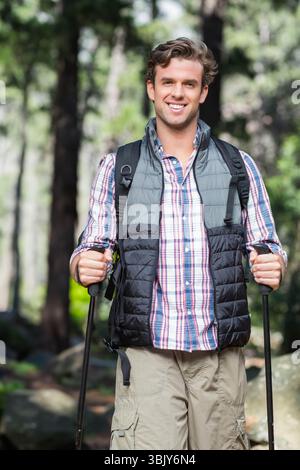 Escursionista maschile che indossa il giubbotto trapuntato sorridente durante il trekking nella pineta con pali e zaino Foto Stock