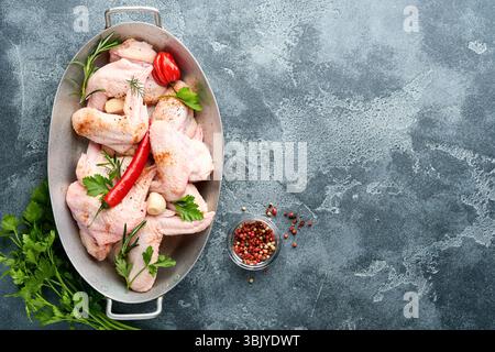 Ali di pollo crude in padella o recipiente di metallo con spezie e ingredienti per cucinare su ardesia grigia scura, pietra o fondo di cemento. Foto Stock