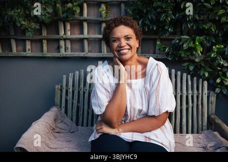 Donna anziana sorridente in casuali che guarda la macchina fotografica mentre si siede fuori nel cortile Foto Stock