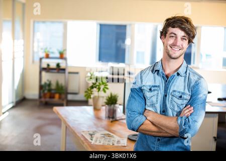 Uomo in piedi armato a croce in uno spazio open Office accanto a una scrivania in legno con computer portatile e piante, spazio fotocopie Foto Stock