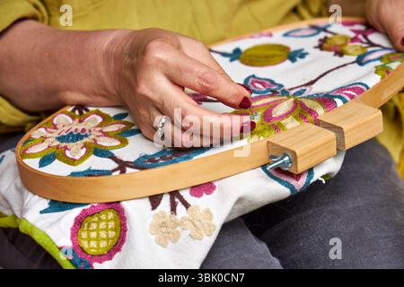 Vista frontale ravvicinata delle mani di una donna che ricama un motivo decorativo su un tessuto allungato in un cerchio da ricamo. Le mani sono concentrate e stabili Foto Stock