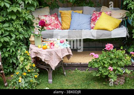 Un accogliente angolo esterno per la creatività in un giardino estivo: Un'altalena imbottita funge da posto rilassante per il ricamo, circondato da una vegetazione lussureggiante e. Foto Stock