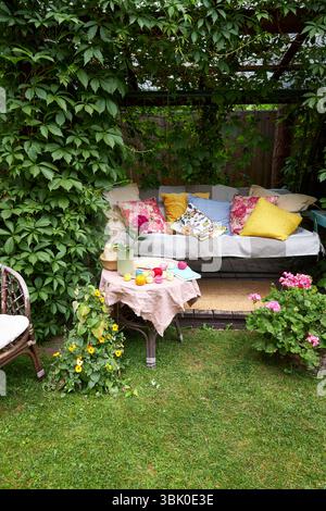 Un accogliente angolo esterno per la creatività in un giardino estivo: Un'altalena imbottita funge da posto rilassante per il ricamo, circondato da una vegetazione lussureggiante e. Foto Stock