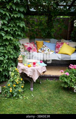 Un accogliente angolo esterno per la creatività in un giardino estivo: Un'altalena imbottita funge da posto rilassante per il ricamo, circondato da una vegetazione lussureggiante e. Foto Stock