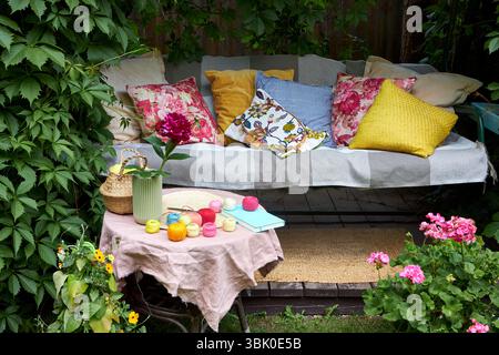Un accogliente angolo esterno per la creatività in un giardino estivo: Un'altalena imbottita funge da posto rilassante per il ricamo, circondato da una vegetazione lussureggiante e. Foto Stock