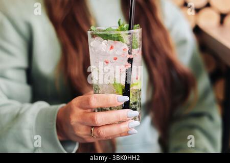 Cocktail mojito fresco in mano alla donna. Drink estivo con menta, ghiaccio e calce in vetro alto. Bevanda rinfrescante con cannuccia. Stile di vita all'aperto. Foto Stock