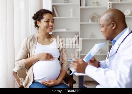 Sorridente donna millennial nera incinta che si consulta con un medico afroamericano maschile Foto Stock