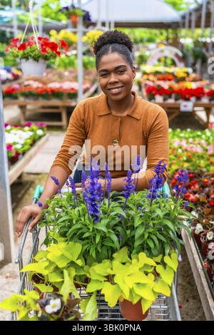Giovane donna felice che spinge un carrello della spesa, seleziona fiori e piante vivaci mentre si diverte in un vivace centro giardino Foto Stock