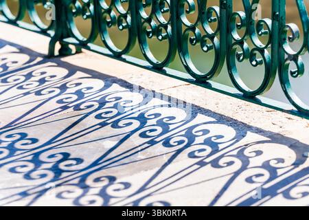 La luce del sole crea intricate ombre di una ringhiera verde in ferro battuto sul passaggio in pietra di un ponte sul fiume. Foto Stock