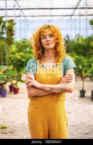Giovane donna con capelli ricci rossi e tatuaggi che indossano tute gialle, in piedi con le braccia incrociate nella serra vivaistica, sembra sicura di sé Foto Stock