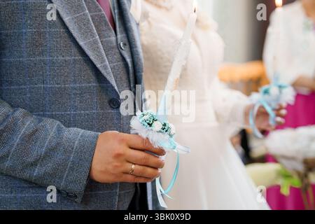 Primo piano di uno sposo con una tuta a plaid grigia che tiene una candela di nozze decorata durante una cerimonia cristiana ortodossa, mostrando un abito bianco e una chiesa Foto Stock