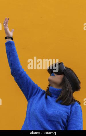 Ritratto verticale isolato di una donna con abiti blu che indossano occhiali vr e toccano qualcosa di invisibile nella realtà virtuale. Sfondo giallo Foto Stock