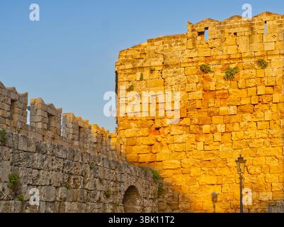 La luce del sole dorata illumina le fortificazioni medievali in pietra nella città vecchia di Rodi con mura merlate e un fossato erboso Foto Stock