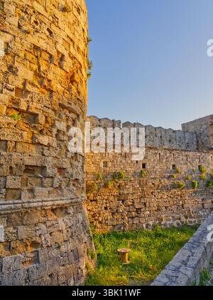 La luce del sole dorata illumina le fortificazioni medievali in pietra nella città vecchia di Rodi con mura merlate e un fossato erboso Foto Stock