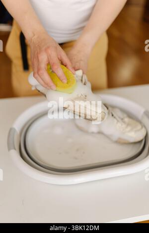 Foto corta verticale di uomo che pulisce le sneakers bianche con schiuma e spazzola, strofinando le scarpe atletiche sporche nel lavabo della casa. Foto Stock