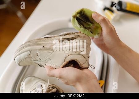 La governante strofina le sneakers bianche sporche con spugna e sapone nel lavabo, primo piano, colpo corto. Concetto di servizio di pulizia e lavori domestici Foto Stock
