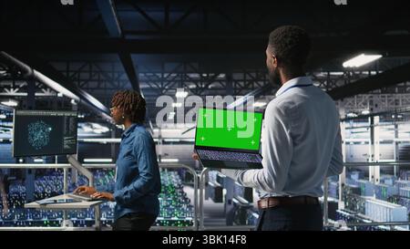 Programmatori di data center su piattaforma industriale che utilizzano notebook con schermo verde e collaborano alle attività. Dipendenti di server hub che visualizzano un'architettura di modelli linguistici di grandi dimensioni utilizzando un notebook e un PC mockup Foto Stock