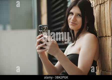 Una giovane donna con capelli castani di media lunghezza sta fuori, sorridendo mentre scatta un selfie. Lo sfondo presenta una parete testurizzata e una luce solare brillante Foto Stock