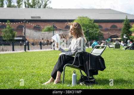 Donna scomoda che si sventola con fogli di carta come un ventilatore mentre si siede sulla sedia all'esterno sul prato. Foto Stock