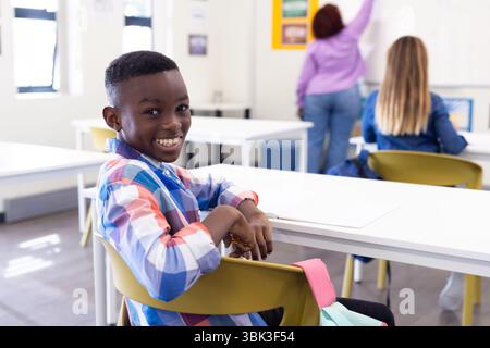 Un ragazzo sorridente in classe seduto alla scrivania mentre un'insegnante donna scrive a bordo, a scuola Foto Stock