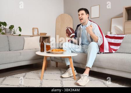 Tifoso sportivo maschile con pallone da rugby e bandiera USA che guarda la partita a casa Foto Stock