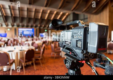 Una videocamera professionale su un treppiede allestito in una spaziosa sala conferenze, con vista su tavoli rotondi e un palco, pronta per registrare un seminario di lavoro o. Foto Stock