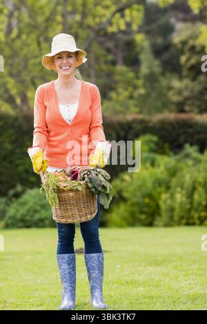 Giardiniere medio adulto che indossa cappello di paglia e guanti sul prato con cesto di carote e verdi Foto Stock