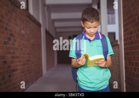 Ragazzo in piedi nel corridoio scolastico con pareti in mattoni che leggono un libro giallo e indossano uno zaino blu Foto Stock