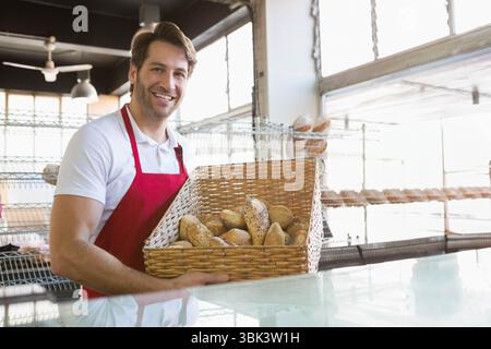 Lavoratore di panetteria maschile che indossa il grembiule rosso sorridente e tiene un cestino di panini dietro il banco di vetro Foto Stock