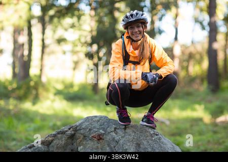 Donna accovacciata sulla roccia nella foresta con giacca arancione, casco da bicicletta e zaino Foto Stock