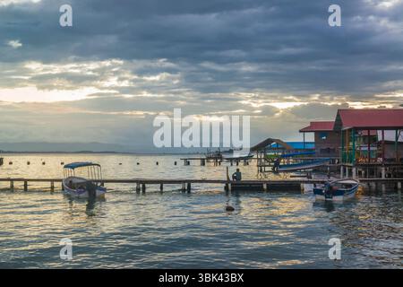 L'uomo guarda l'acqua su un molo di legno con barche ormeggiate al tramonto. Old Bank, Isla Bastimentos, Bocas del Toro, Panama Foto Stock