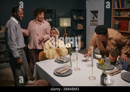 I membri della famiglia sorridono e chiacchierano mentre si radunano intorno al tavolo della cena in un ambiente accogliente, mostrando calore e gioia. Set da tavolo con piatti, bicchieri e candele Foto Stock