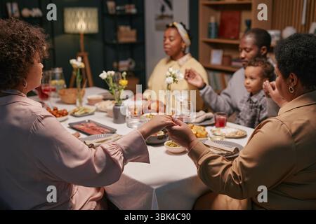 Colpo ritagliato di membri della famiglia nera che si tengono per mano seduti intorno al tavolo in preghiera prima della cena di gala nella sala da pranzo Foto Stock