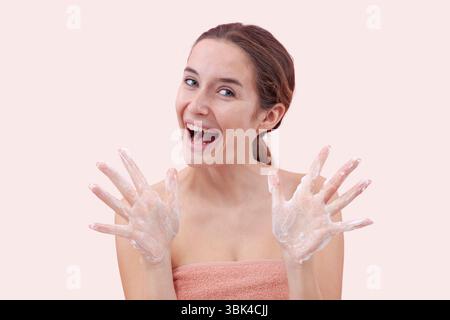 Giovane donna felice con detergente schiumoso sulle mani, tenendoli aperti verso la fotocamera prima di lavarsi il viso. Routine per la cura della pelle per una pulizia, una salute Foto Stock