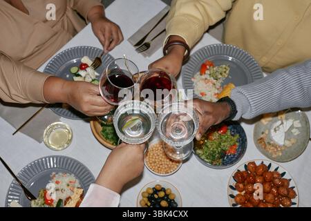Foto ad alto angolo di diversi gruppi di persone che alzano i bicchieri intorno al tavolo pieni di piatti e bevande, per festeggiare le vacanze con la famiglia e gli amici a casa Foto Stock
