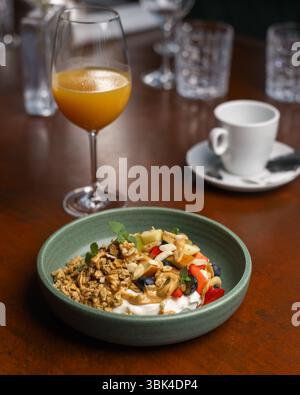 Una ciotola di yogurt ricoperta di muesli, frutta e noci e un bicchiere di succo su un tavolo di legno durante la colazione. Foto Stock