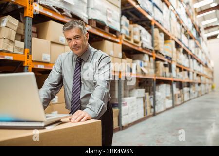 Uomo adulto maturo che si appoggia alle scatole e controlla il computer portatile in magazzino con scaffali in metallo e spazio di copia Foto Stock